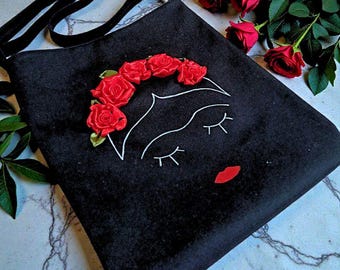 May include: A black shoulder bag featuring a white embroidered outline of a face with closed eyes and a red fabric lip accent. The bag is decorated with a crown of five dimensional red fabric roses across the top of the embroidered forehead.