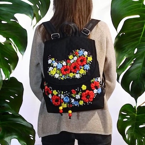 Könnte beinhalten: Ein schwarzer Rucksack mit lebendiger Bandstickerei aus roten Mohnblumen, gelben Blüten und kleinen blauen Blumen. Der Rucksack verfügt über einen Tragegriff, verstellbare Schultergurte, einen Kordelzug mit roten und gelben Perlen.