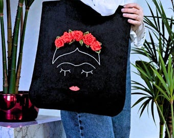 May include: A black velvet tote bag featuring a white embroidered outline of a face with closed eyes and a pink lip detail. The design is accented with a crown of three-dimensional red fabric roses positioned above the embroidered forehead.