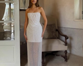 Può includere: Un abito bianco senza spalline con intricate perline e una gonna trasparente con frange. Il corpetto presenta un motivo dettagliato di perle, mentre la gonna scende fino al pavimento. Questo elegante abito è adatto per eventi formali.