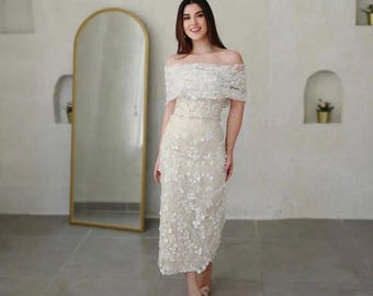 Puede incluir: Vestido color crema, hasta la rodilla, con adornos florales. El vestido presenta un escote de hombros descubiertos y un corpiño ajustado. Los detalles florales descienden por la falda. Un espejo dorado se ve al fondo.