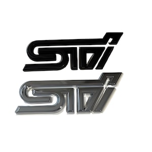 May include: Two STI emblems are shown, one black and one chrome, on a white backdrop. The emblems feature a stylized design with the letters "STI" in a bold, angular font. The black emblem is glossy, while the chrome one has a reflective finish.