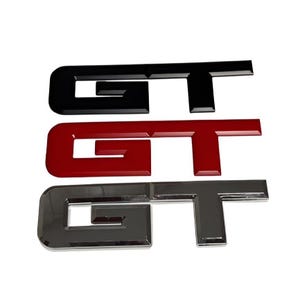 May include: Three GT emblem badges in black, red, and chrome. The emblems are glossy and have a three-dimensional appearance. The letters are stylized and have a modern font. The badges are likely for automotive use.