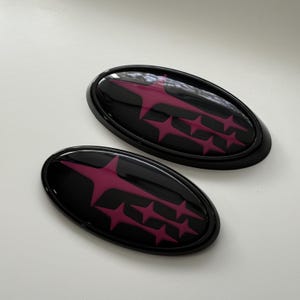 May include: Two black oval car emblems with a glossy finish. Each emblem features a stylized magenta starburst design. The emblems are set against a white background, highlighting their shape and color.