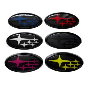 May include: Six oval-shaped automotive emblems featuring the Subaru star cluster logo. Each emblem has a black background with a glossy finish. The star designs are colored in blue, red, white, black, pink, and yellow, respectively.