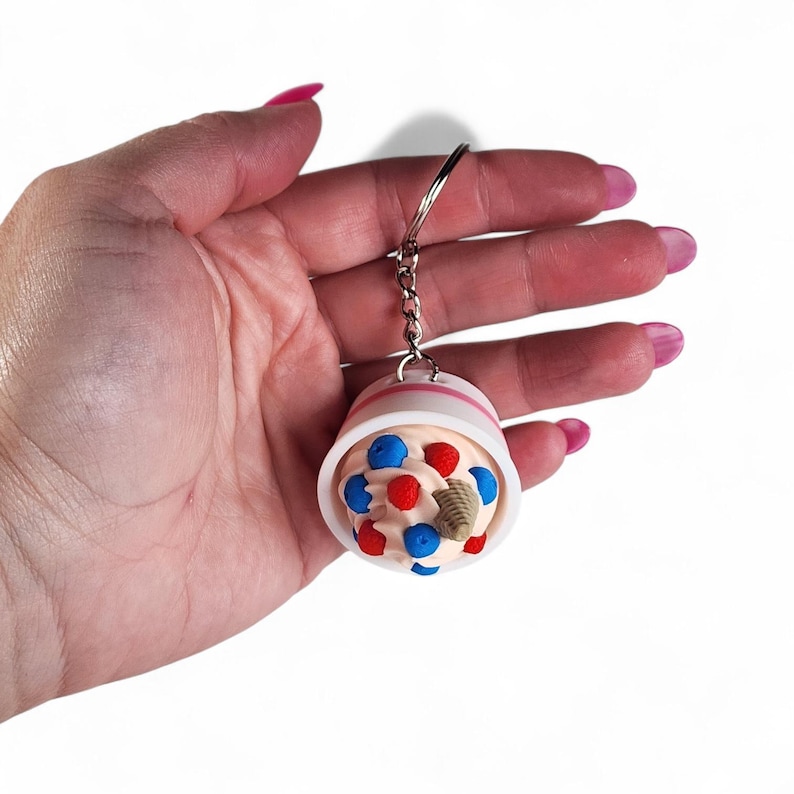 Op de afbeelding: Een miniatuur ijscoupe sleutelhanger. De ijscoupe zit in een witte beker met een roze streep en is bedekt met rode en blauwe hagelslag en een klein wafelkegeltje. De sleutelhanger is bevestigd aan een zilveren ketting.