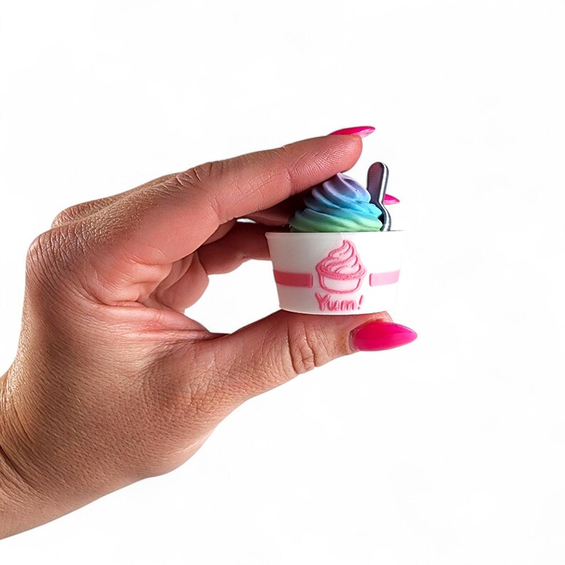 May include: A miniature white cup of colorful soft-serve ice cream with a spoon. The cup has a pink stripe and a pink ice cream cone graphic with the word "Yum!" in pink. The ice cream is a swirl of blue, green, and purple.