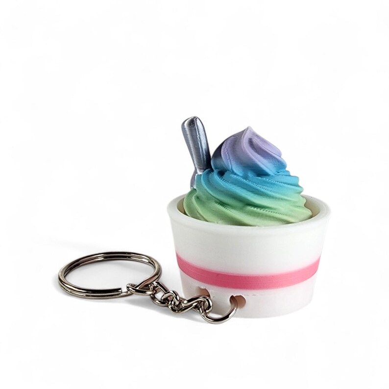 May include: A whimsical keychain featuring a miniature ice cream sundae. The sundae has a swirl of colorful, rainbow-colored ice cream in a white cup with a pink stripe. A small silver spoon is inserted into the ice cream. The keychain has a silver ring and chain.