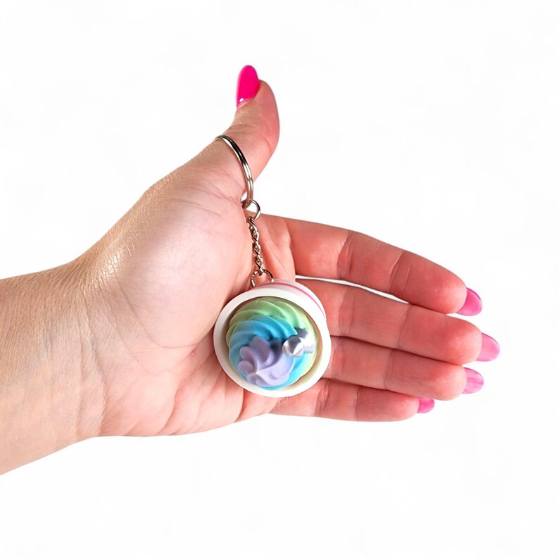 May include: A round keychain with a colorful, swirled design in shades of blue, green, and purple, resembling a miniature dessert. The keychain has a silver-colored ring and chain. A small, silver-colored embellishment is visible on the front.