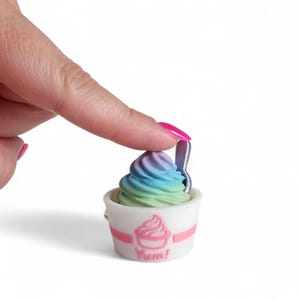 May include: A miniature ice cream cup with a swirl of pastel-colored ice cream. The cup is white with a pink stripe and a pink ice cream cone graphic with the word "Yum!" below. A small silver spoon is inserted into the ice cream.