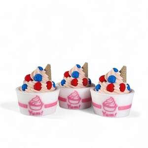 Op de afbeelding: Drie miniatuur ijsdesserts in witte bekers met roze accenten. Elk heeft een werveling van roze softijs, rode en blauwe bessen en een kleine wafel. De bekers hebben een roze ijslogo en het woord "Yum!"