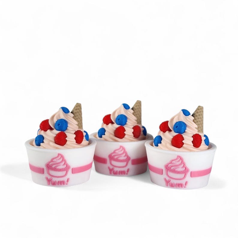 Op de afbeelding: Drie miniatuur ijsdesserts in witte bekers met roze accenten. Elk heeft een werveling van roze softijs, rode en blauwe bessen en een kleine wafel. De bekers hebben een roze ijslogo en het woord "Yum!"