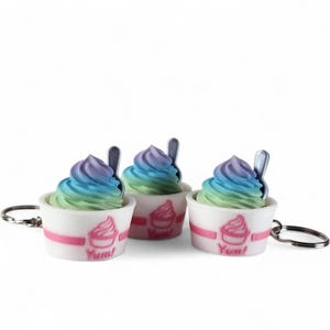 May include: Three ice cream keychains with rainbow-colored swirls, each in a white cup with a pink band and the word "Yum!" The keychains have a small spoon and a silver keyring. The ice cream is a pastel mix of blue, green, and purple.
