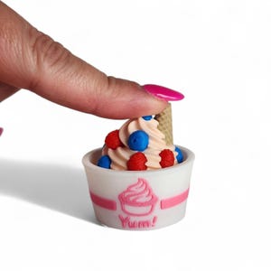 Op de afbeelding: Een miniatuur ijsdessert in een witte beker met een roze streep en het woord "Yum!". Het ijs heeft een werveling van lichtroze softijs, gegarneerd met rode frambozen en blauwe bosbessen, en een kleine wafel.