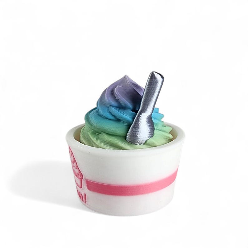 May include: A colorful dessert in a white cup with a pink stripe. The dessert has a swirl of green, blue, and purple. A silver spoon is inserted into the dessert. The cup has a pink graphic on the side.
