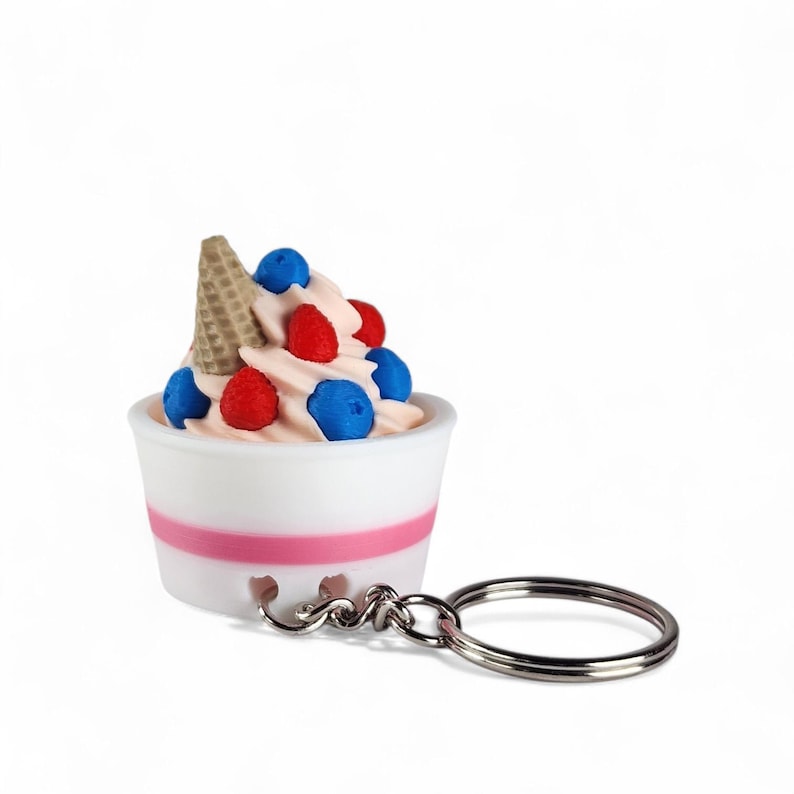 Op de afbeelding: Een novelty sleutelhanger met een miniatuur ijscoupe. De ijscoupe heeft roze softijs, gegarneerd met rode en blauwe bessen en een klein wafelhoorntje. De ijscoupe zit in een witte beker met een roze streep en is bevestigd aan een zilveren sleutelhanger.