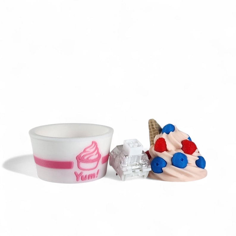 Op de afbeelding: Een witte beker met een roze streep en een roze ijsje met de tekst "Yum!" naast een doorzichtig, rechthoekig object en een softijsje met rode en blauwe bessen.