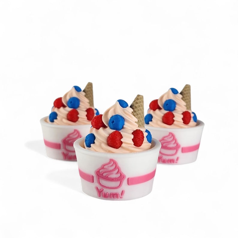 Op de afbeelding: Drie bekers softijs met roze wervelingen, gegarneerd met rode frambozen, blauwe bosbessen en een klein wafelkegeltje. De bekers zijn wit met een roze streep en het woord "Yum!"
