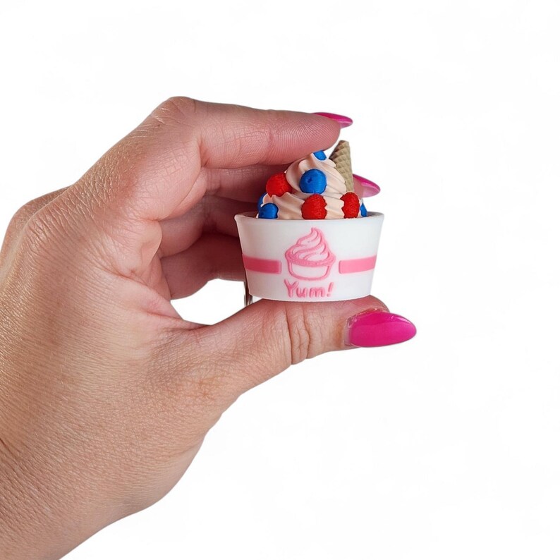 Op de afbeelding: Een miniatuur softijs dessert in een witte beker met roze strepen en een roze ijsje afbeelding. Het ijs is gegarneerd met rode en blauwe bessen en een klein wafelkegeltje. Het woord "Yum!" staat op de beker.