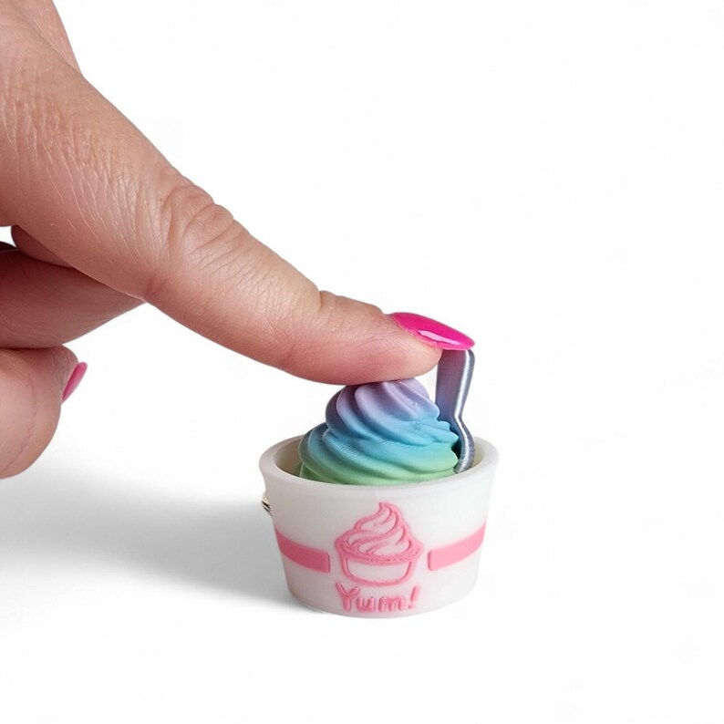 May include: A miniature white ice cream cup with a pink band and a pink ice cream cone graphic. The cup contains a swirl of colorful ice cream in shades of blue, green, and purple. A small silver spoon is inserted into the ice cream. The word "Yum!" is printed on the cup.