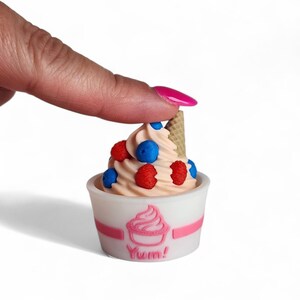 Op de afbeelding: Een miniatuur ijscoupe in een witte beker met een roze streep en het woord "Yum!". Het ijs is perzikkleurig, gegarneerd met blauwe en rode bessen en een klein wafelhoorntje.