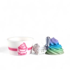 May include: A white paper cup with a pink band and a pink ice cream graphic, the word "Yum!" is below the graphic. Next to the cup are a clear, rectangular object, a silver spatula, and a colorful swirl of ice cream.