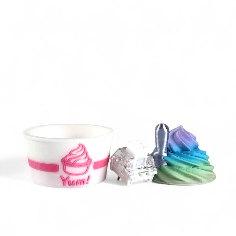 May include: A white paper cup with a pink band and a pink ice cream graphic, the word "Yum!" is below the graphic. Next to the cup are a clear, rectangular object, a silver spatula, and a colorful swirl of ice cream.