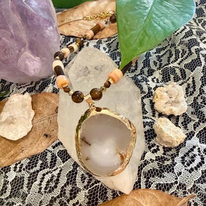 Può includere: Una collana con un grande ciondolo in pietra bianca irregolare con un bordo metallico dorato. La catena è composta da perline cilindriche alternate color pesca e marrone scuro, intervallate da perline rotonde marroni più piccole e distanziatori dorati.