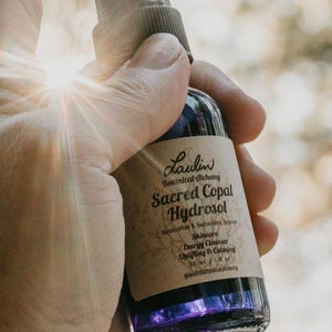 May include: A glass bottle with a purple liquid inside. The bottle has a brown label with the text "Laulin Botanical Alchemy Sacred Copal Hydrosol Handcrafted & Sustainable Sourced Skincare Energy Cleanser Uplifting & Calming 30 ml/1 fl oz Laulinbotanicalalchemy".
