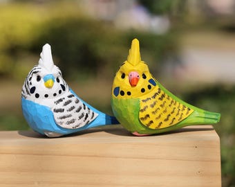 Puede incluir: Dos figuritas de pájaros de madera, una azul y blanca, la otra verde y amarilla, con rayas negras y amarillas. Los pájaros están posados ​​en una superficie de madera.