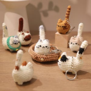 May include: A collection of seven crocheted cat keychains in various colors, including white, brown, and gray. The keychains are all in different poses, some sitting, some standing, and some lying down. The keychains are all made with a soft, fuzzy yarn and have cute, expressive faces.
