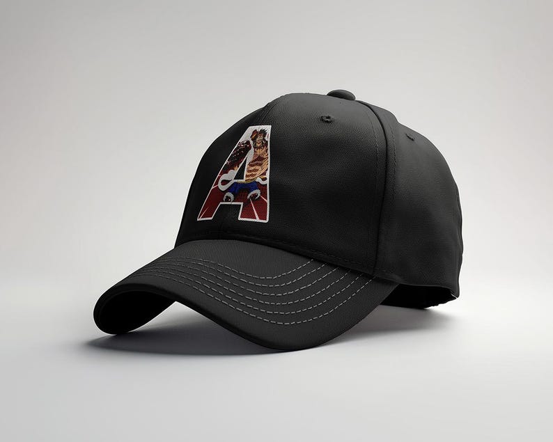 May include: Black baseball cap with a curved brim and a stitched design. The cap features a colorful graphic of a character inside a white and red letter "A". The cap is a stylish accessory.