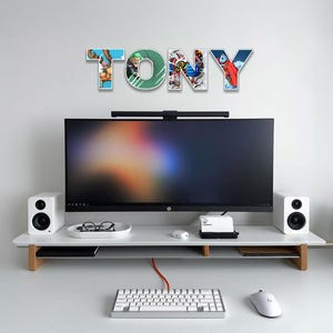 May include: A modern desk setup featuring a large computer monitor, white speakers, and a keyboard. Above the monitor, the name "TONY" is spelled out in colorful, illustrated letters. The desk is white with a wooden support.