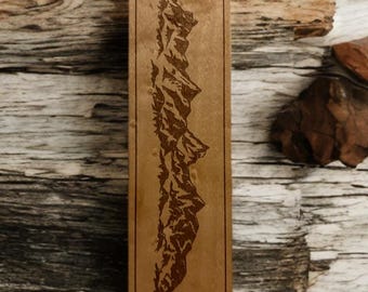 Könnte beinhalten: Ein rechteckiger Holz-Lesezeichen mit einem eingravierten Bergketten-Design. Das Lesezeichen ist hellbraun mit einem dunkelbraunen Rand und einem Loch für eine Quaste. Der Hintergrund ist eine Holzoberfläche, ca. 15 cm lang.