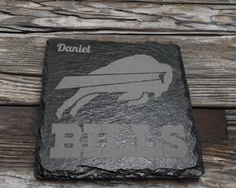 May include: A square, dark gray slate coaster with the name "Daniel" etched at the top. The Buffalo Bills logo and the word "BILLS" are etched in the center. The coaster has a rough, textured surface.