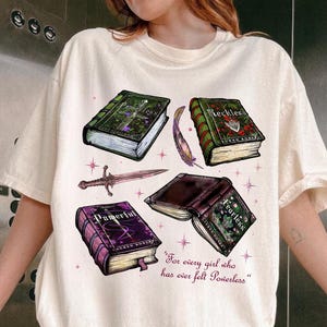 Może przedstawiać: Biały t-shirt z grafiką czterech książek z różnymi tytułami: "Powerful", "Fearless", "Reckless" i "Powerful". Tekst "For every girl who has ever felt powerless" jest nadrukowany pod książkami.