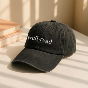 May include: A dark grey baseball cap with the embroidered text "well-read" in white. The cap features a curved peak and a classic six-panel design. The hat is on a white surface.