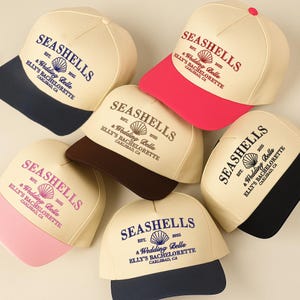 Pode incluir: Uma coleção de bonés de beisebol com o texto "SEASHELLS" e "Elly's Bachelorette" impresso na frente. Os bonés têm uma coroa creme e uma aba colorida, com cores como azul marinho, rosa, preto e castanho. Os bonés são para uma despedida de solteira.