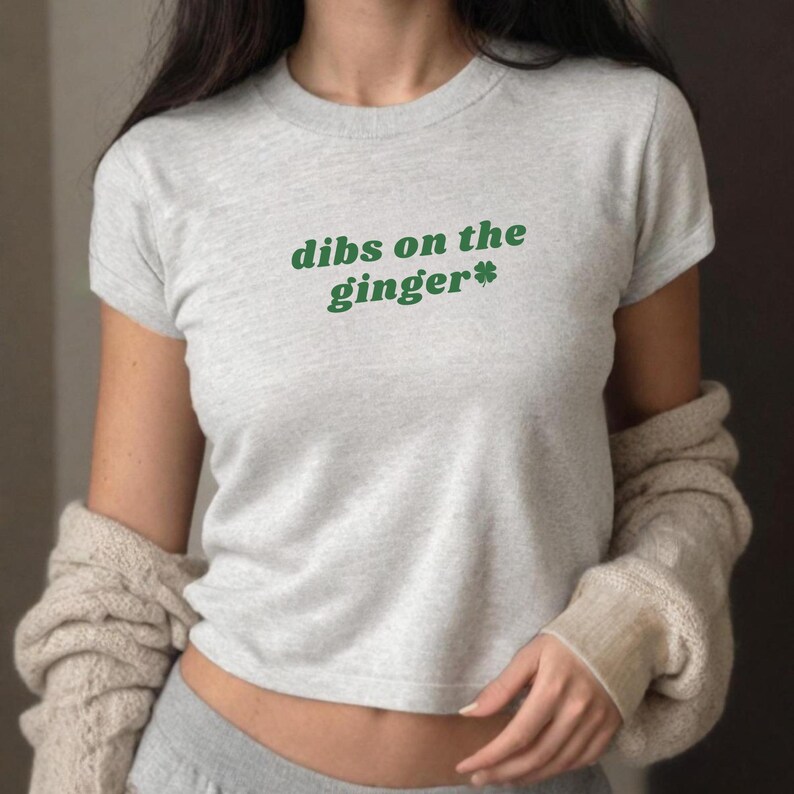 Puede incluir: Una camiseta gris de manga corta con el texto "dibs on the ginger*" en letras verdes. Un tr&eacute;bol de cuatro hojas verde est&aacute; al lado de la palabra "ginger".