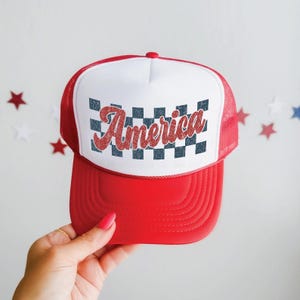 May include: Red and white trucker hat with a red brim and mesh back. The front panel is white with a checkered pattern and the word "America" in red script. A patriotic accessory.