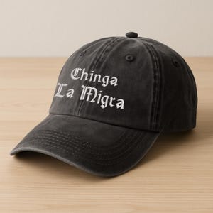 Può includere: Un cappellino da baseball grigio scuro con la scritta ricamata "Chinga La Migra" in lettere gotiche bianche. Il cappellino ha una visiera curva e un bottone sulla parte superiore. Il cappello è realizzato in un materiale morbido e usurato.