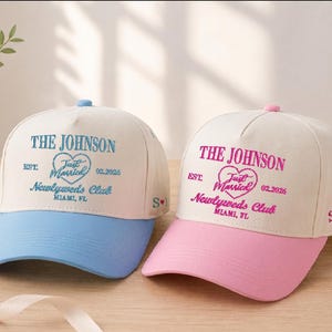 Puede incluir: Dos gorras de béisbol con el texto "THE JOHNSON" y "Just Married Newlyweds Club" bordado en la parte delantera. Una gorra tiene una visera azul, la otra una visera rosa. Ambas tienen las iniciales "S M" bordadas en el lateral.