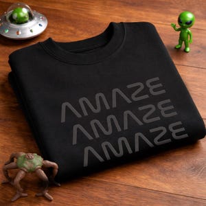 Könnte beinhalten: Schwarzes Sweatshirt mit dem Wort "AMAZE" dreimal in hellgrauer Schrift. Das Kleidungsstück liegt gefaltet auf einer Holzoberfläche. Spielzeug-Alienfiguren und ein Raumschiffmodell sind ebenfalls zu sehen.