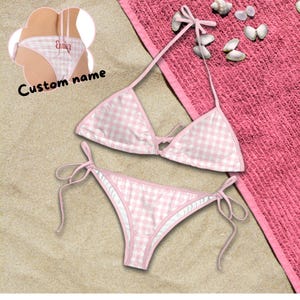 May include: A pink and white gingham bikini set with tie closures. The bikini top and bottom are displayed on a sandy surface next to a pink towel and seashells. The words "Custom name" are visible.