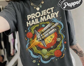 Op de afbeelding: Donkergrijs t-shirt met de tekst "PROJECT HAIL MARY" en een kleurrijke afbeelding van een ruimteschip in de ruimte. Het ontwerp bevat planeten en een wervelend melkwegstelsel. Het shirt heeft korte mouwen en een ronde hals.