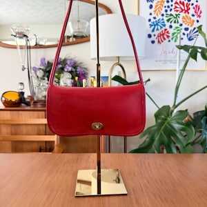 May include: A rectangular red leather shoulder bag featuring a slim profile, a single thin shoulder strap, and a centered gold-tone metal turn-lock closure on the front flap. The bag is suspended on a gold-colored metal display stand.