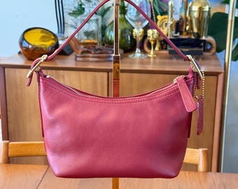 Könnte beinhalten: Eine kleine, halbmondförmige Handtasche aus glattem, dunkelrotem Leder. Sie verfügt über einen einzelnen Tragegriff, der mit goldfarbenen Metallbeschlägen befestigt ist, einen Reißverschluss oben und einen passenden Lederanhänger mit einer goldfarbenen Kugelkette.