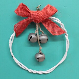 May include: A white wire wreath with a red burlap bow and silver bells hanging from it. The wreath is on a turquoise background.
