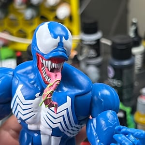 Puede incluir: Figura de acción azul de Venom, con detalles blancos y un emblema de araña blanco en el pecho. La figura tiene una boca grande con dientes afilados y una larga lengua rosa. La figura está en una pose dinámica.