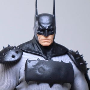 May include: A Batman action figure with a black cowl and pointed ears. The figure has a gray and black costume with a bat symbol on the chest and spiked shoulder armor. The figure's face is visible, with a stern expression.